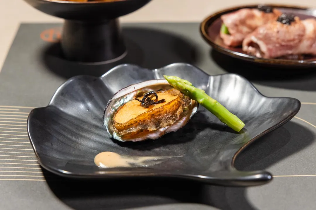 台北旭集三間門市推出期間限定料理〈嫩煎鮑魚佐蕈菇奶油醬〉，主廚先將飽滿厚實的鮑魚煎至表層些微酥香，點綴蕈菇奶油白醬，再佐以昆布鹽及新鮮蘆筍，甘甜鹹鮮交織。（圖/饗賓集團提供）